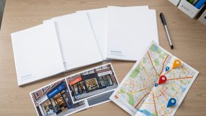 Espace de travail organisé avec plusieurs dossiers professionnels, photographies de devantures, carte urbaine et marqueurs de localisation réorganisés après le déménagement de plusieurs agences.